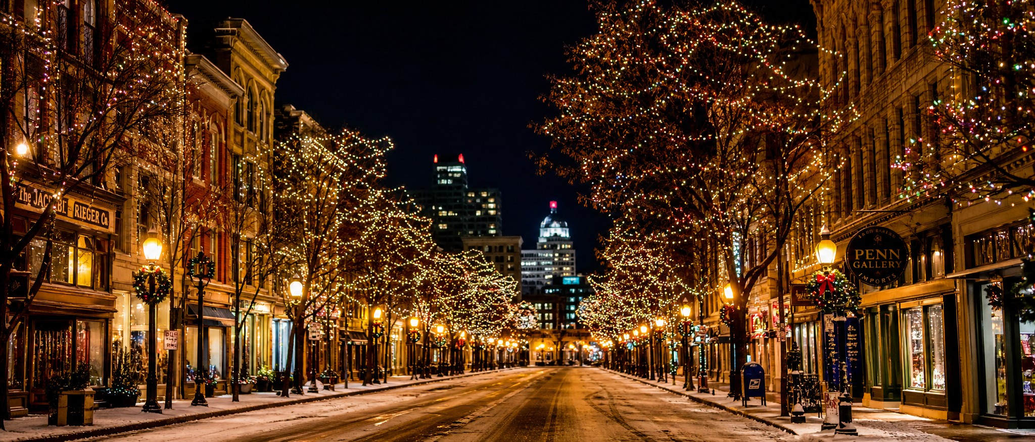 A charming Pittsburgh city street with trees and buildings decorated in warm white and colorful Christmas lights at night